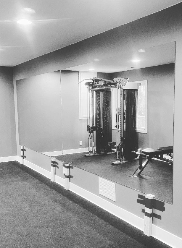 Home gym with large mirror, workout equipment including a pulley machine, adjustable bench, and open windows in a room with carpeted floor.