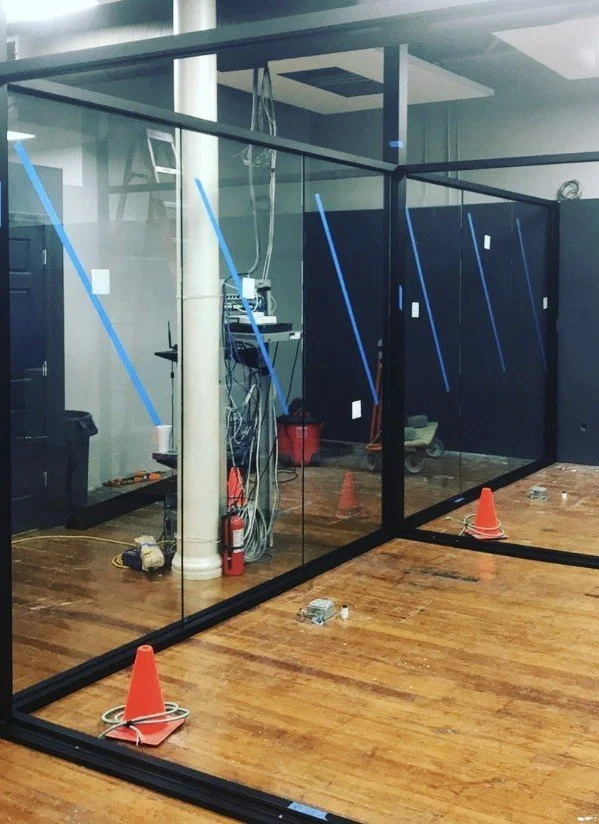 Interior of a room under construction with glass walls, orange traffic cones, and various tools and equipment on the floor.