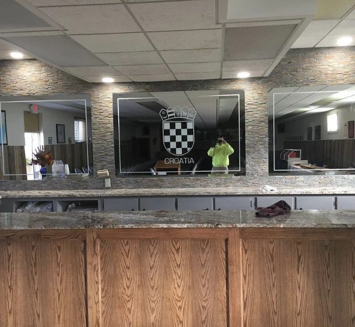 Mirror with Croatian soccer team logo, reflecting a person in a bright green jacket taking the photo. The wall has brick decor, and above the counter, there are three ceiling lights.
