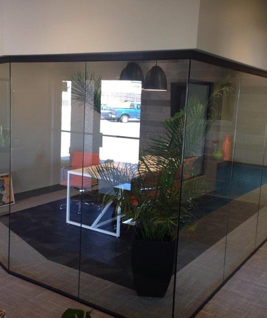 Modern office meeting room with glass walls, a white table, black chair, orange couch, large green potted plant, and pendant lights.