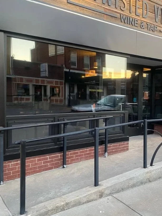 Reflective glass window of a restaurant or store with a metal railing outside and a brick wall base, showing reflections of nearby buildings and a parked car.