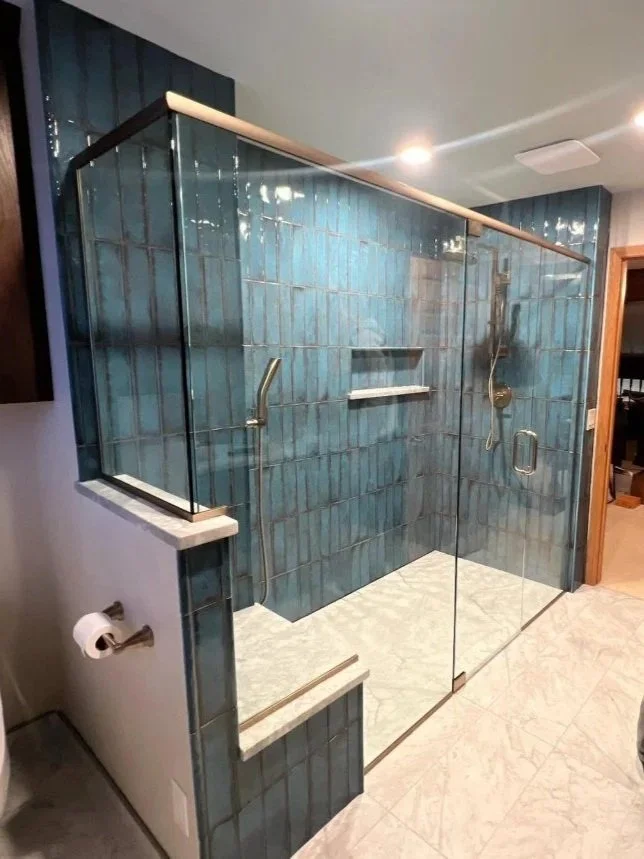 A walk-in shower with greenish-blue tiles, glass walls, a built-in seat, and a bathroom with white marble flooring.