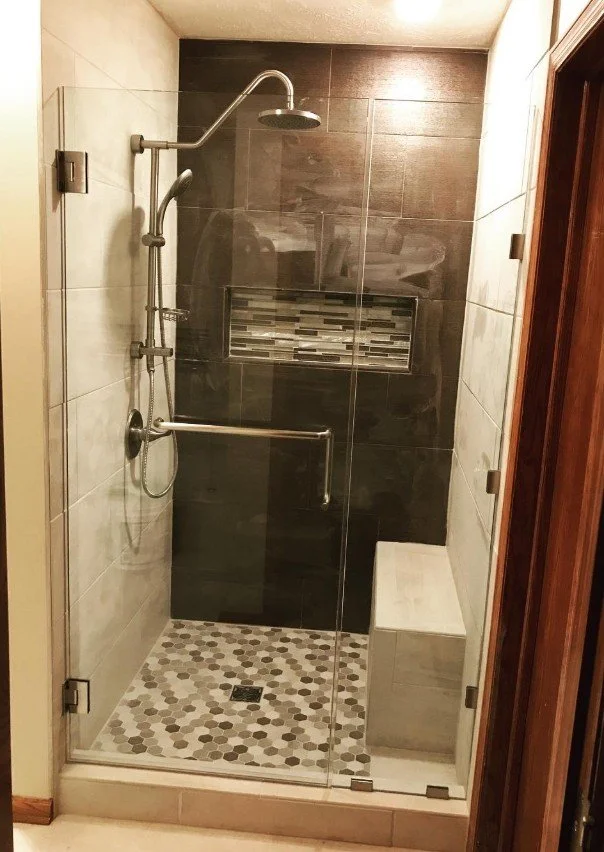 A walk-in shower with a glass door, black and beige tiles, a built-in bench, and a tiled wall niche for toiletries.
