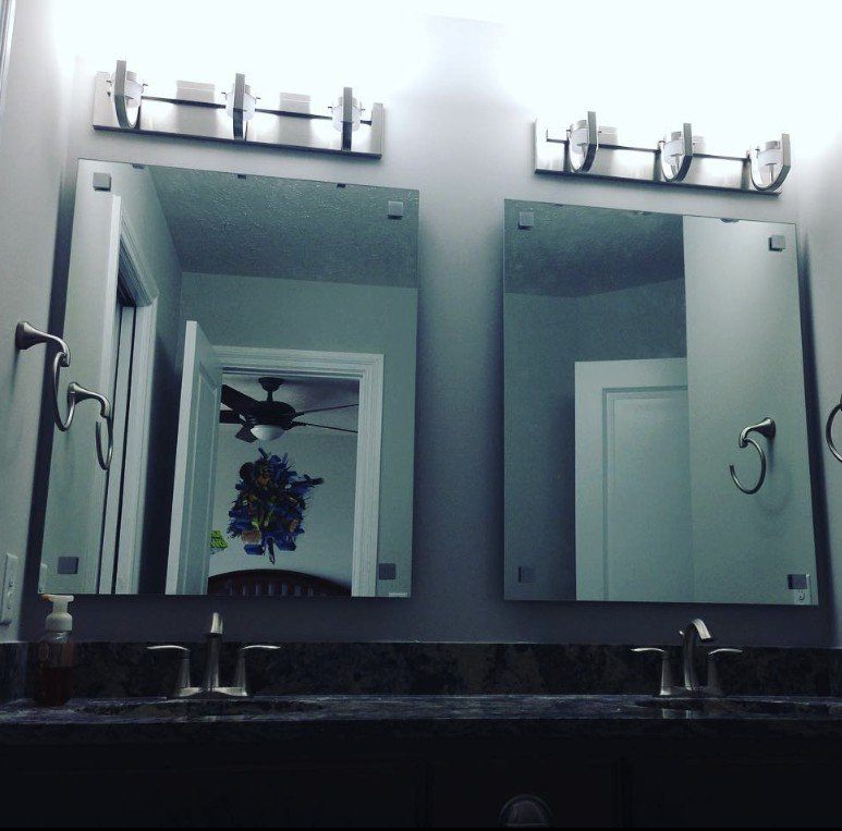 Two bathroom mirrors above a dark granite countertop with two sinks, with a reflection showing a ceiling fan and a doorway with a colorful wall hanging.