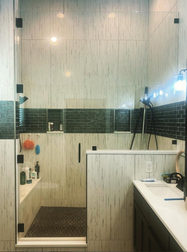 Modern bathroom with a partitioned walk-in shower, black lower tiles, white upper tiles with subtle veining, black vanity, and various toiletries on a white countertop.