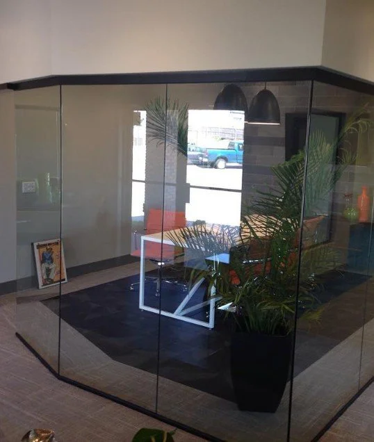 Indoor modern office or lounge area with glass walls, a black rug, a white table, and orange seating. Large potted plants and a view of parked cars outside.