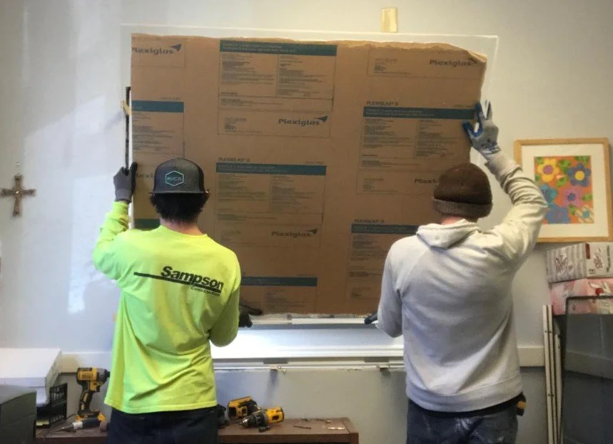 Two men installing a large pane of Plexiglas into a window frame inside a room.