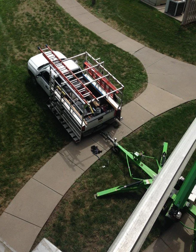 A white utility truck parked on a sidewalk with an extensive ladder rack on the bed, and a green boom lift nearby. The scene is on a grassy area with sidewalks and some buildings.