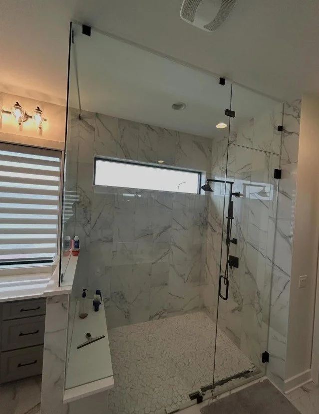 Modern walk-in shower with glass enclosure, marble tile walls, and a long horizontal window. The shower features a rainfall showerhead and handle fixtures. To the left, there is a countertop with bottles of shampoo and soap, and a window with a strip