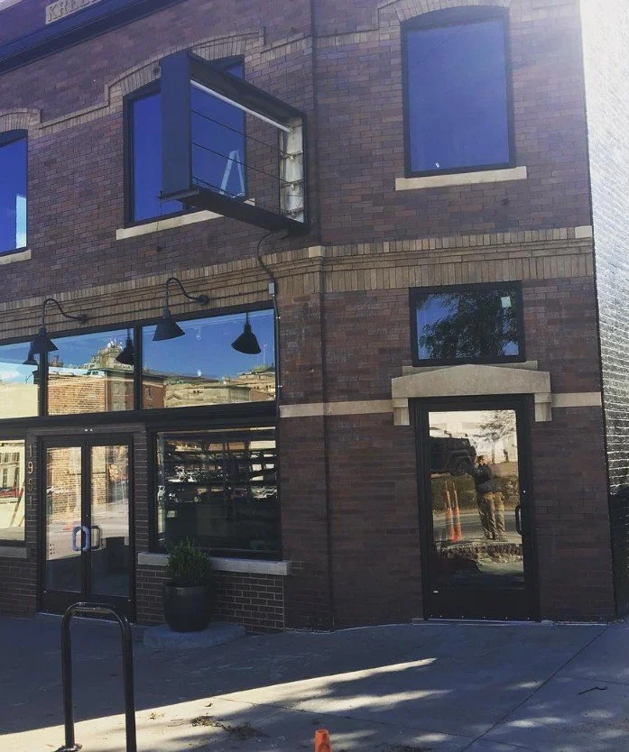 Brick building with large glass windows and a glass door, reflecting the street scene. There is an open window shade on the upper floor, black outdoor wall lights, a potted plant near the entrance, and a person taking the photo reflected in the door.