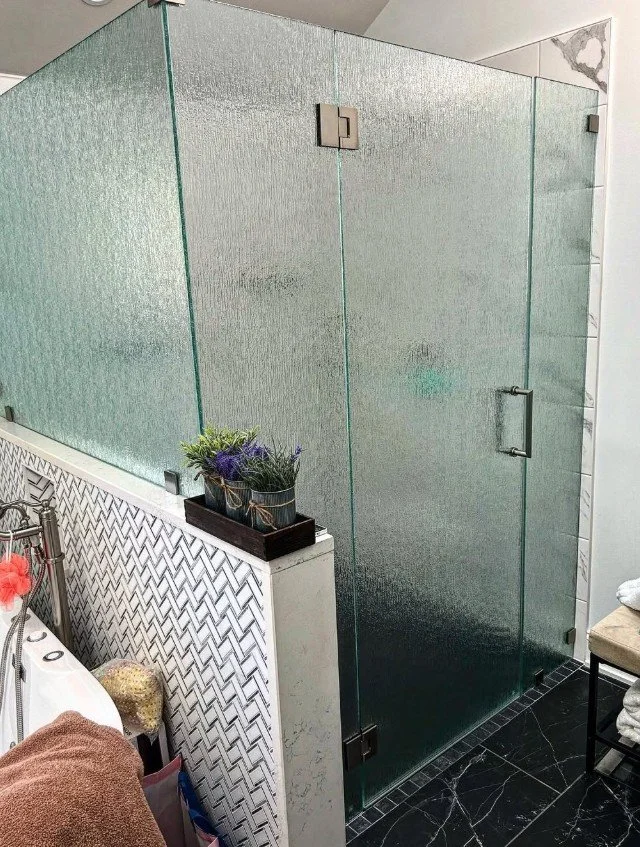 Frosted glass shower stall in a bathroom with a black marble floor, a small plant on a shelf, and towels on a nearby chair.