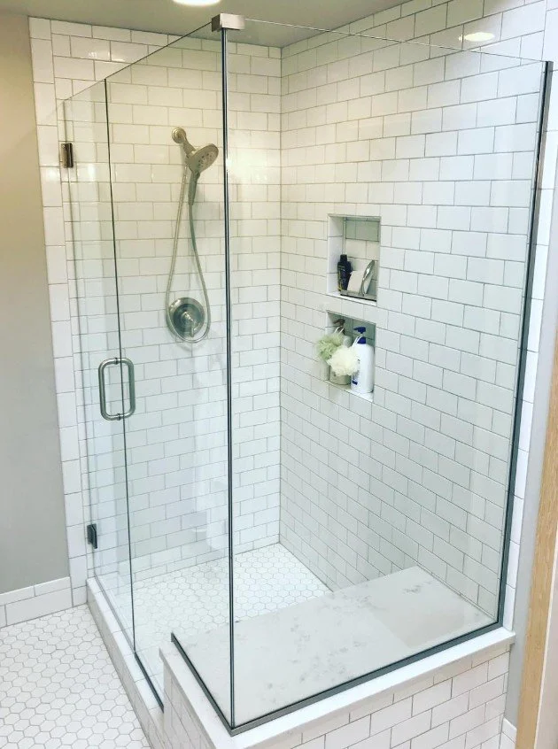 A modern walk-in shower with clear glass walls, white subway tile walls, a built-in shelf with toiletries, a handheld showerhead, and a white hexagonal tile floor.