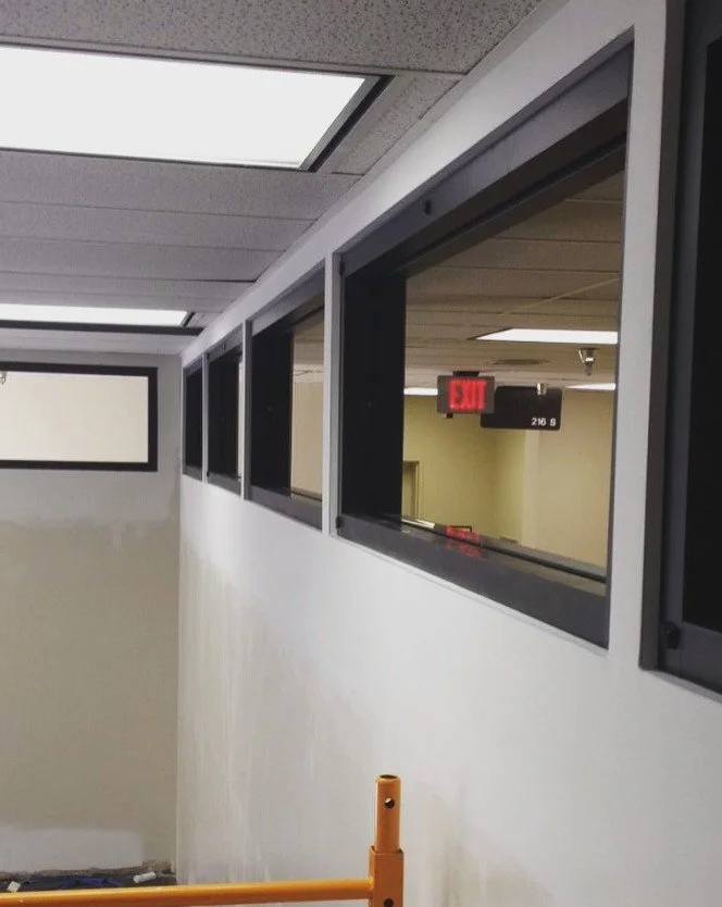 Interior view of a building with a white wall and multiple high rectangular windows. An exit sign and a black room number sign are visible in the background.