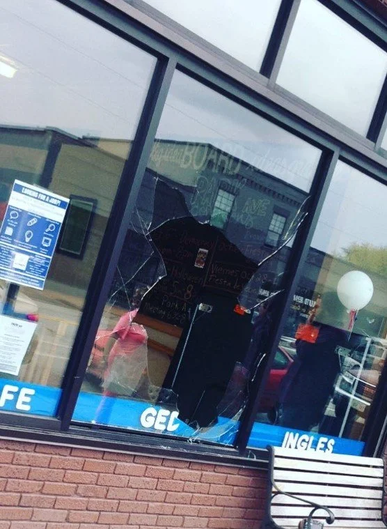 A storefront window with a broken glass panel in the center. The glass is shattered and cracked around the damage. Inside, there is a person in a pink shirt. Outside, there are reflections of cars and people, and a white balloon. The window has signs