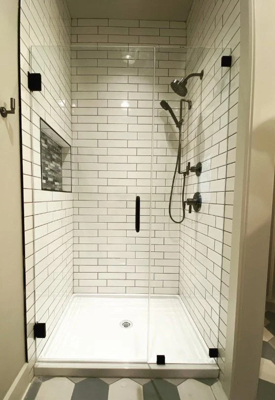 A modern walk-in shower with white subway tiles, a built-in tiled niche, black fixtures, and a glass door. The shower has a rainfall showerhead and a handheld showerhead.