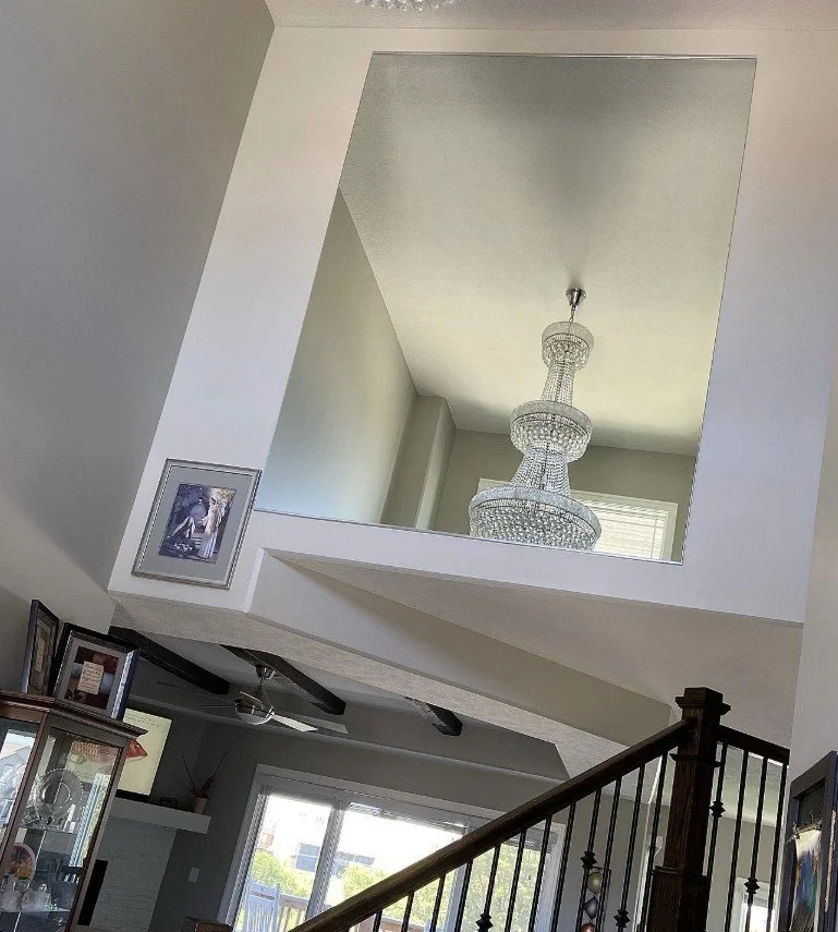 Interior view of a living space with a staircase, high ceiling, and a large window. A crystal chandelier hangs from the upper level, reflecting light.