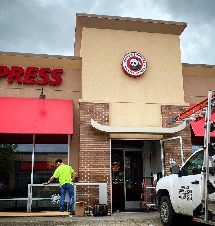 Exterior of a Panda Express restaurant under renovation, with a worker installing a glass panel, a white utility vehicle with a ladder nearby, and the Panda Express logo visible above the entrance.