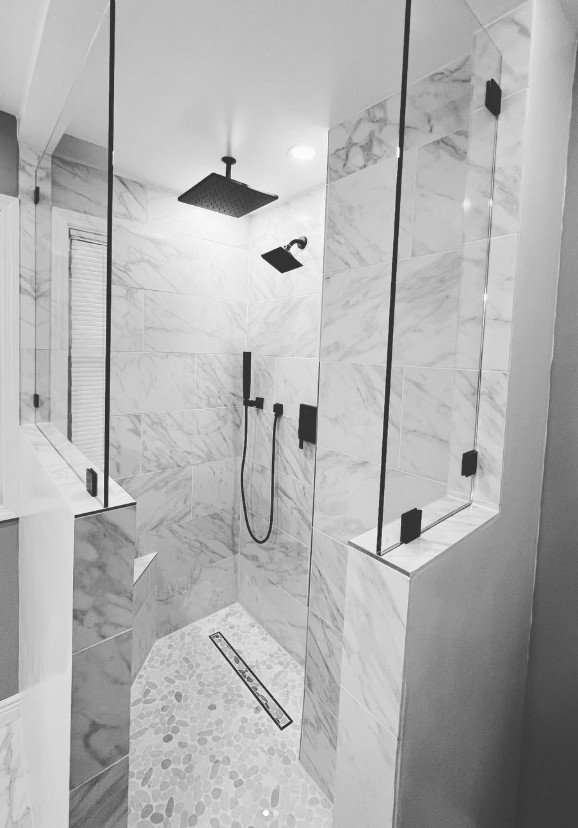 Modern glass-enclosed walk-in shower with marble tiles, rainfall showerhead, handheld shower, and pebble stone floor.