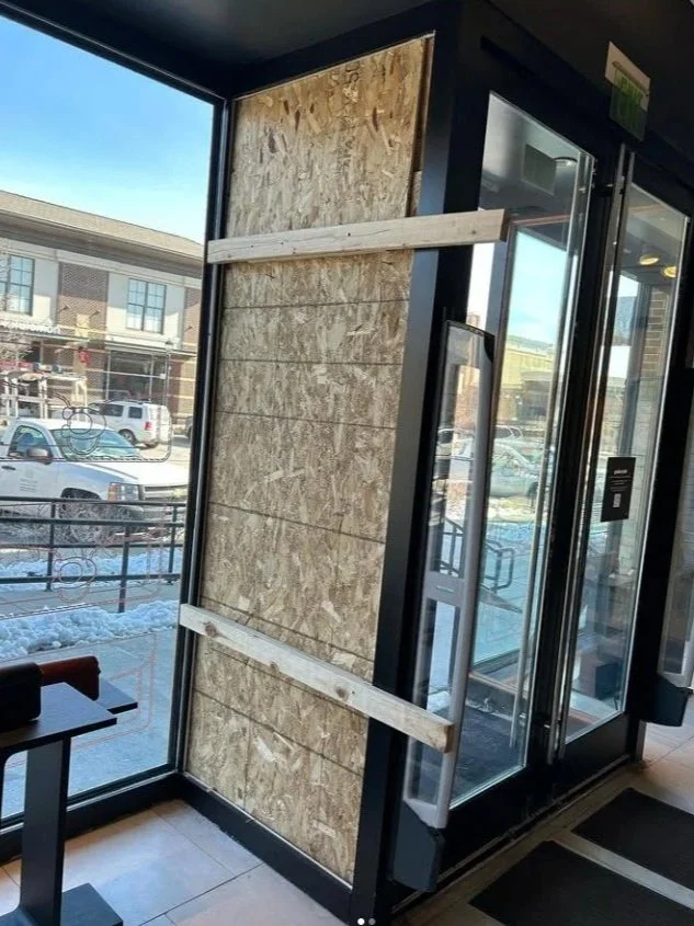 Boarded-up window in a commercial building, with wood panels and horizontal braces, partially covering the glass window.