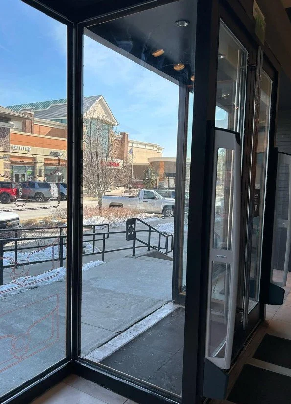 View of a shopping plaza seen through the glass door and window of a store, with parked cars and a snow-covered sidewalk outside.