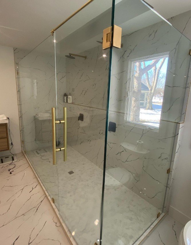 A modern walk-in shower with glass enclosure featuring gold hardware, marble walls and floor, and a window with a view of trees outside.