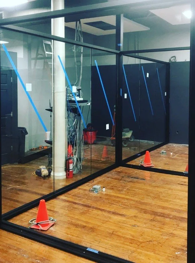 Glass wall partition under construction in a room with a hardwood floor, orange traffic cones, and various tools and wiring.