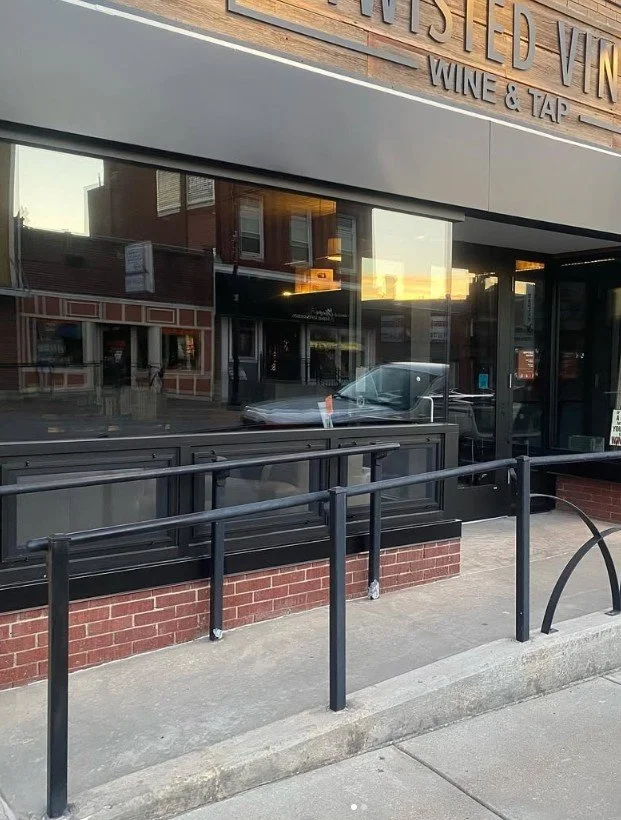 A sidewalk outside a restaurant with large glass windows reflecting buildings and a parked car, a black railing runs along the sidewalk, and the sign reads "WINE & TAP".