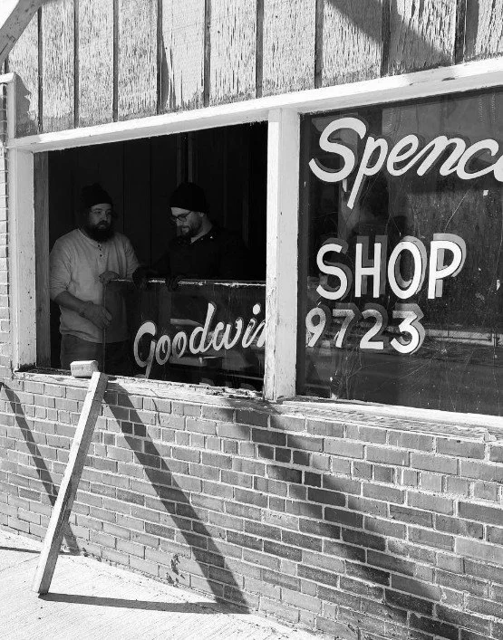 Two men in beanies and glasses inside a storefront with large glass windows, one wearing a light shirt and the other a dark shirt, standing near a counter. The storefront has signs that read 'Spenc,' 'Shop,' 'Goodwi,' and a partial phone number '9723