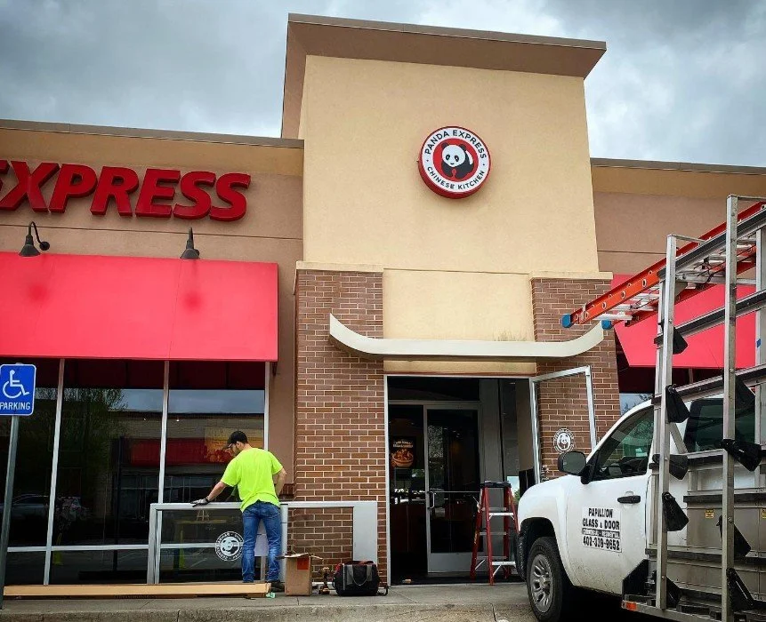 Outside of a Panda Express restaurant with a red awning and signage. A worker in a neon yellow shirt is setting up or cleaning outside. A white utility truck with a ladder is parked nearby. The building has a brick facade with beige walls and a circular panda logo.