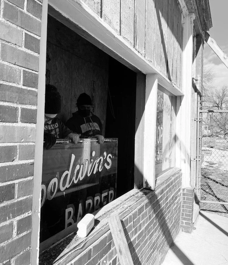 Three people inside a barber shop looking out through a large glass window. Their faces are mostly obscured, and they are wearing masks and winter hats. The exterior of the shop is brick, and there is a sign that reads 'Dodvina's Barber' in the windo