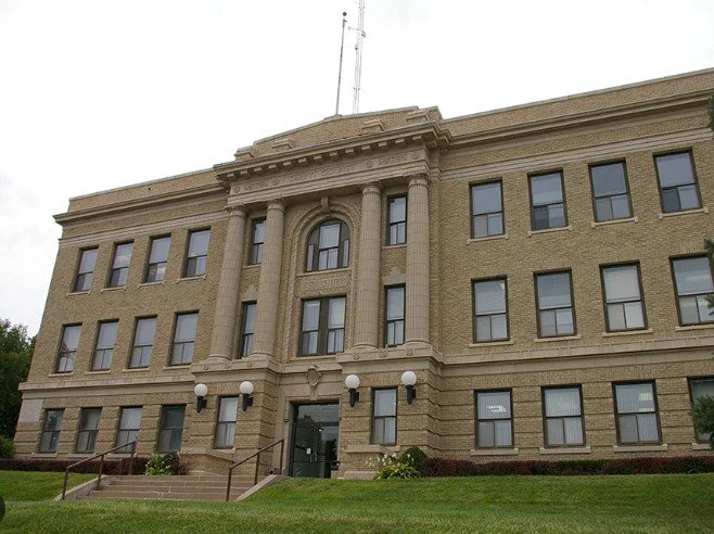 Front view of a beige brick government or administrative building with columns above the entrance, stairs, and a landscaped lawn.