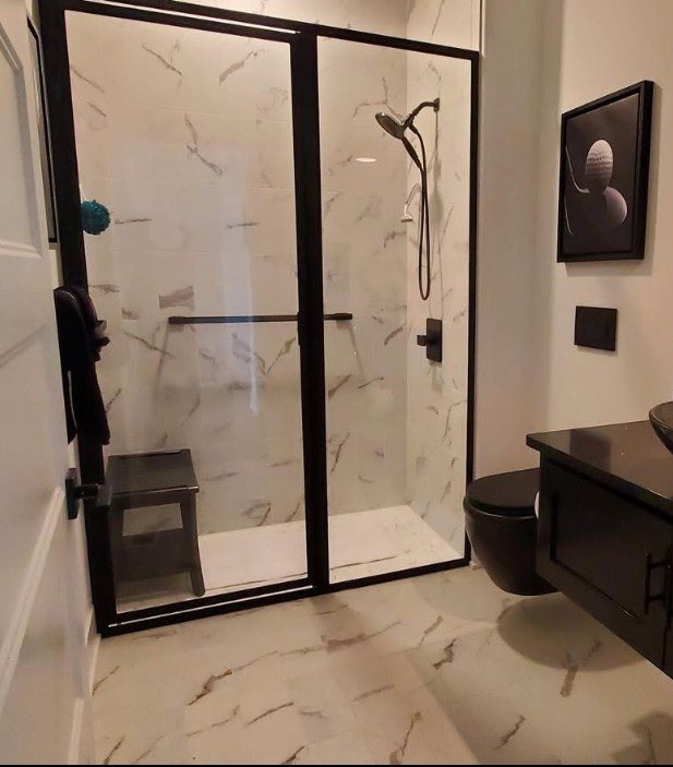 Modern bathroom with a glass shower enclosure, marble walls and floor, black fixtures, a wall art piece, and a black vanity with a black stool inside the shower.