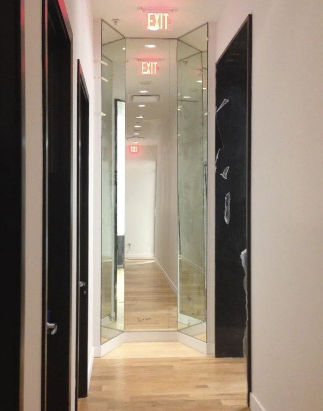 A narrow hallway with wooden flooring, black and white walls, and a mirror at the end, with illuminated exit signs above.