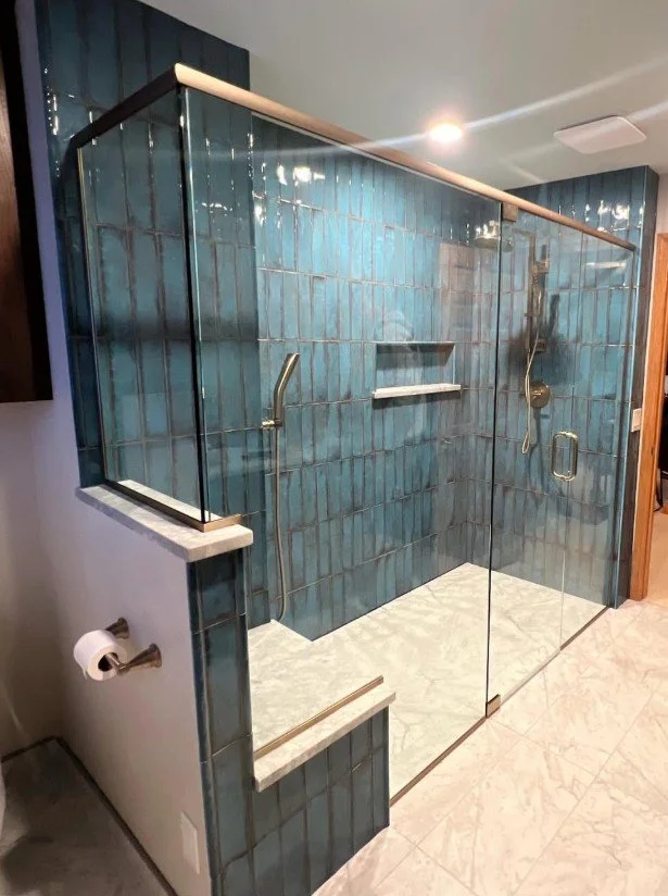 Glass-enclosed walk-in shower with blue tiling, built-in shelf, and two metal grab bars, located in a bathroom with tiled flooring.