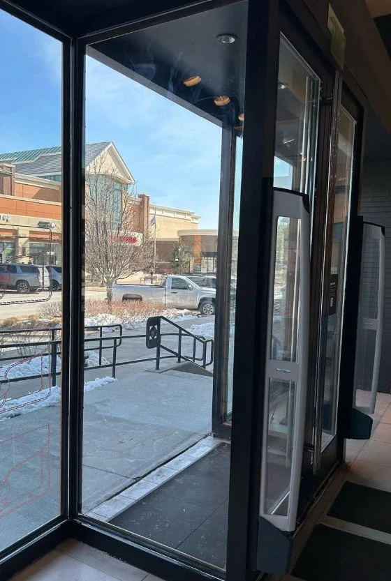 View from inside a building looking outside through large glass windows and door. Outside is a sidewalk, a railing, snow on the ground, parked cars, and a shopping center with retail stores.