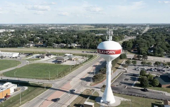 A water tower with the word 'ELKHORN' painted on its side, situated near a street and surrounded by various buildings and parking lots in a suburban area.