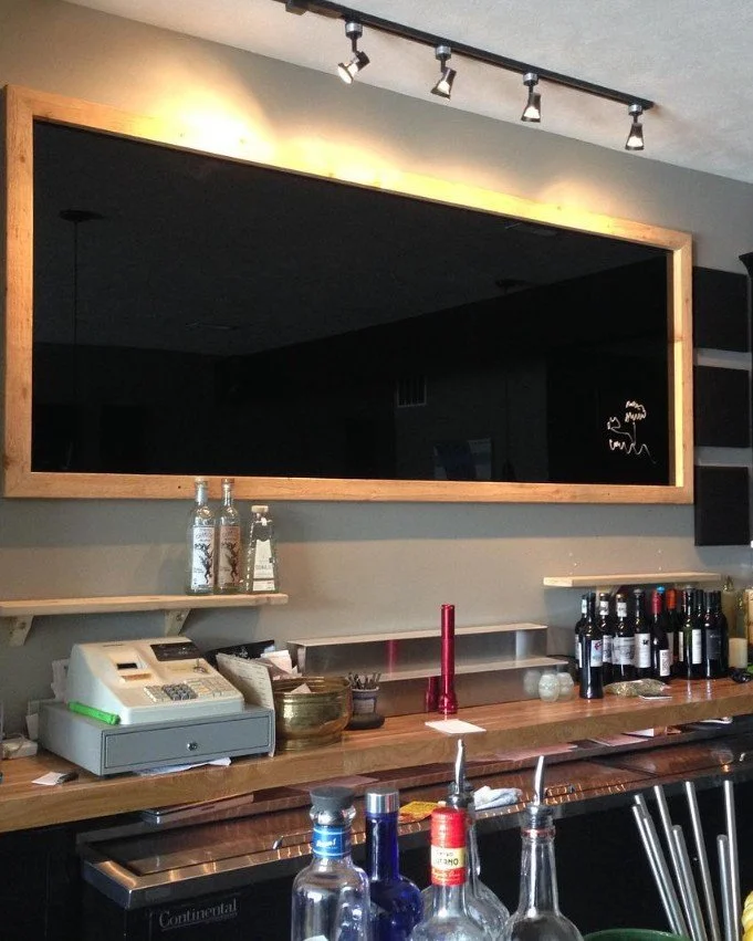 A bar counter with bottles of alcohol, a cash register, and glasses. A large mirror with a wooden frame hangs on the wall above the bar, reflecting an empty and dark room. Track lighting is mounted on the ceiling above the mirror.
