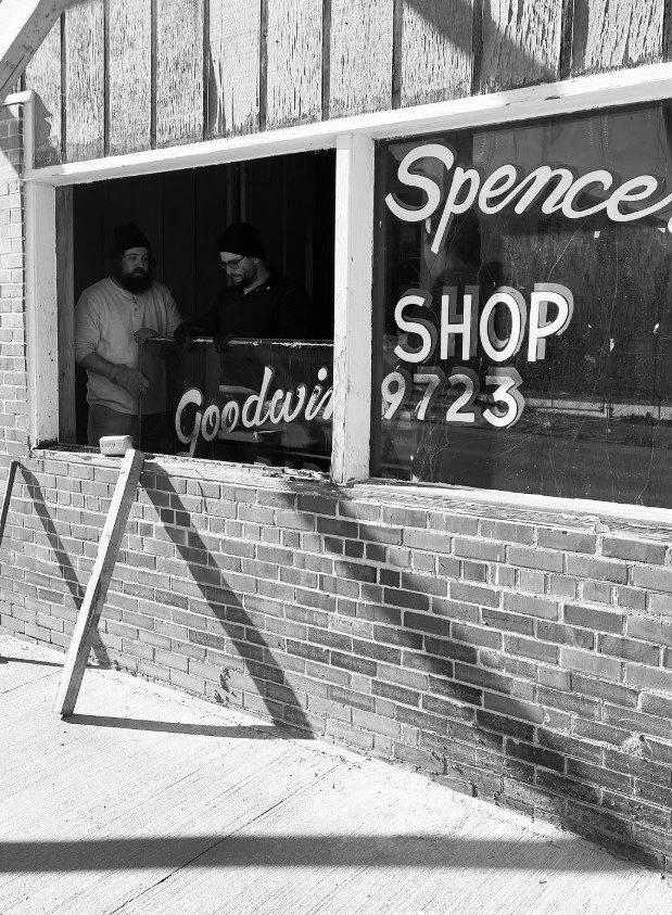 Two men standing inside a shop, seen through a large window in a brick building, with signage for "Spence Shop" and "Goodwin" visible on the window.