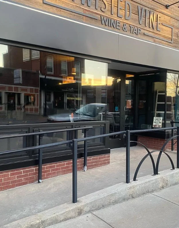 Exterior of a wine bar called 'Wisted Vino' with a black railing and large glass windows, reflecting nearby buildings and a parked silver car.