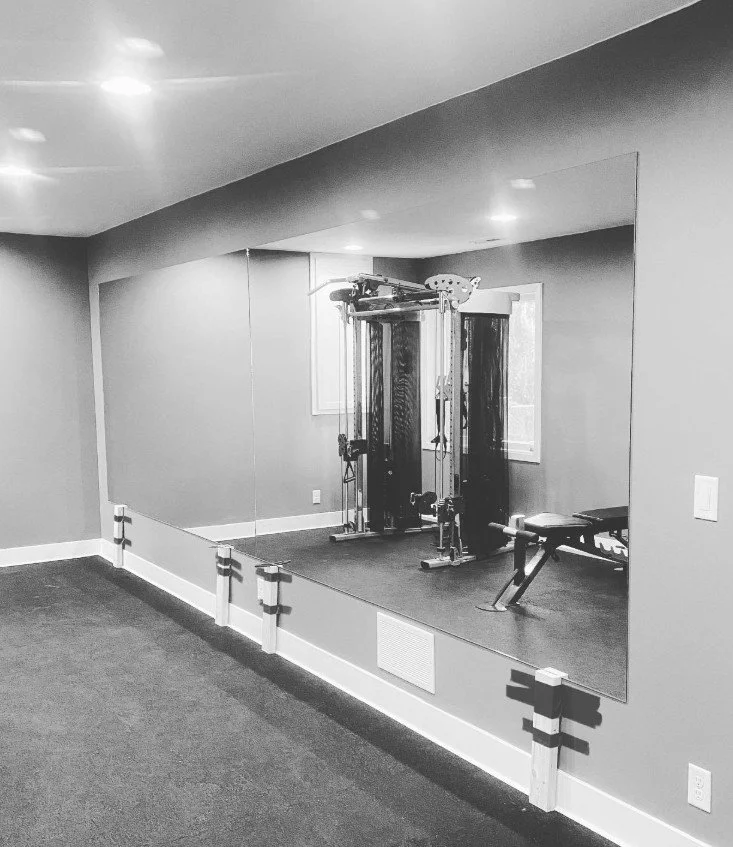 A home gym with a cable machine and adjustable workout bench viewed through a large mirror, with a window on the wall.