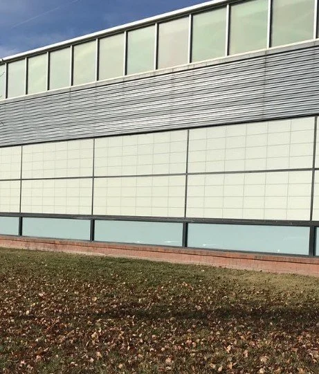 Close-up of a modern building's exterior wall, featuring large glass windows at the top, horizontal metal siding in the middle, and divided glass panels near the ground level, with a grassy area in front.