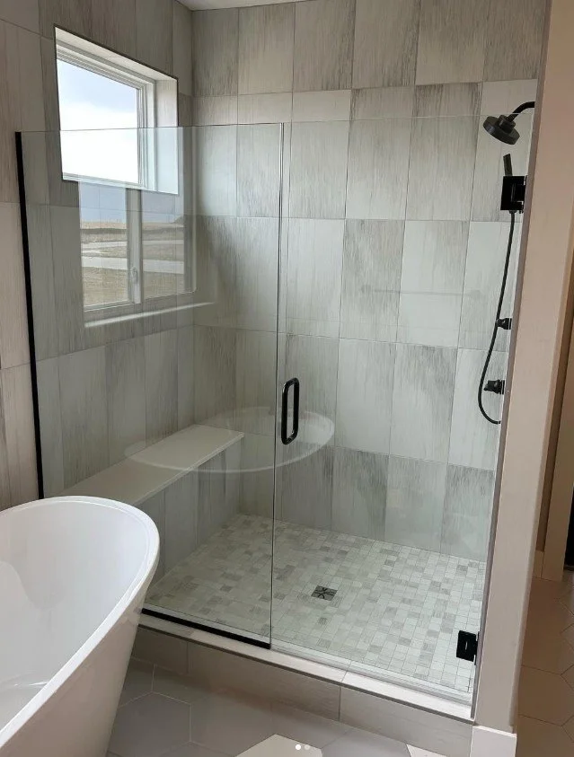 A modern bathroom with a walk-in shower featuring beige tiled walls and a glass door. The shower has a black showerhead and a built-in bench, with a small window providing natural light.
