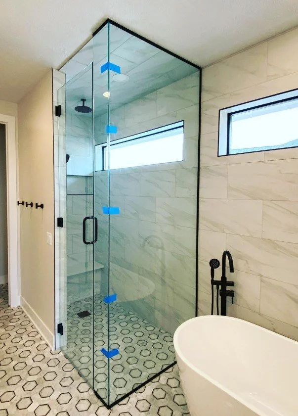 A modern bathroom featuring a glass-enclosed shower with black fixtures, marble-patterned wall tiles, two horizontal narrow windows, a freestanding bathtub, and geometric hexagonal floor tiles.