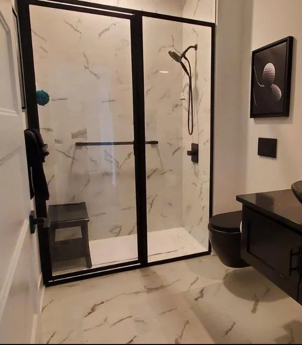 Modern bathroom with a glass shower enclosure, marble walls and floor, black fixtures, a toilet, and a black vanity with a framed space-themed picture on the wall.