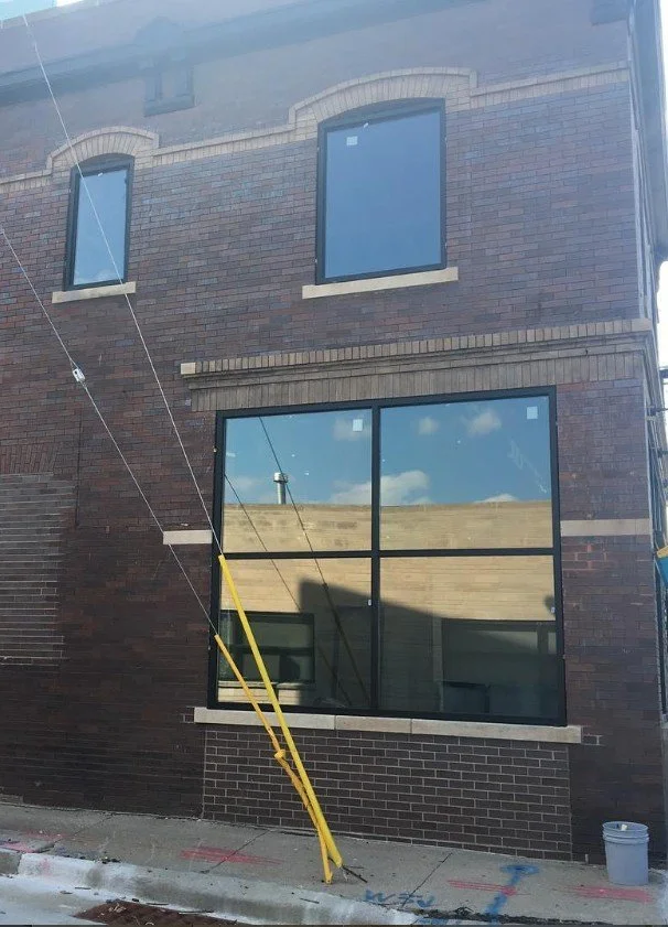 A three-story building with brick exterior and large front windows reflecting a blue sky with clouds. There is a yellow pole leaning against a rope or wire attached to the building, possibly related to construction or maintenance work.
