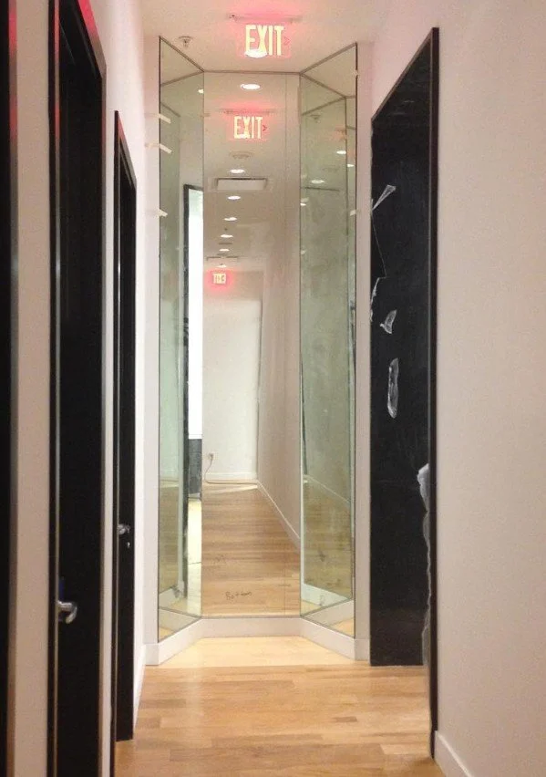 A narrow hallway with light wooden floors, white walls, and black accents, featuring a large mirror at the end with an exit sign above and reflections of ceiling lights.