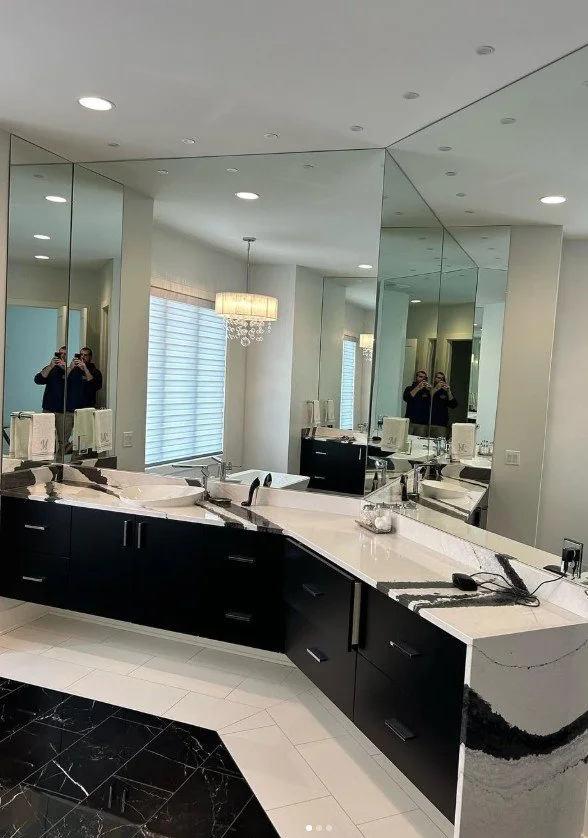 Modern bathroom with black and white marble countertops, large mirrors, and a chandelier, with two people taking a photo reflected in the mirrors.