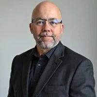 A bald man with glasses wearing a black suit jacket and black shirt, standing against a plain light gray background.