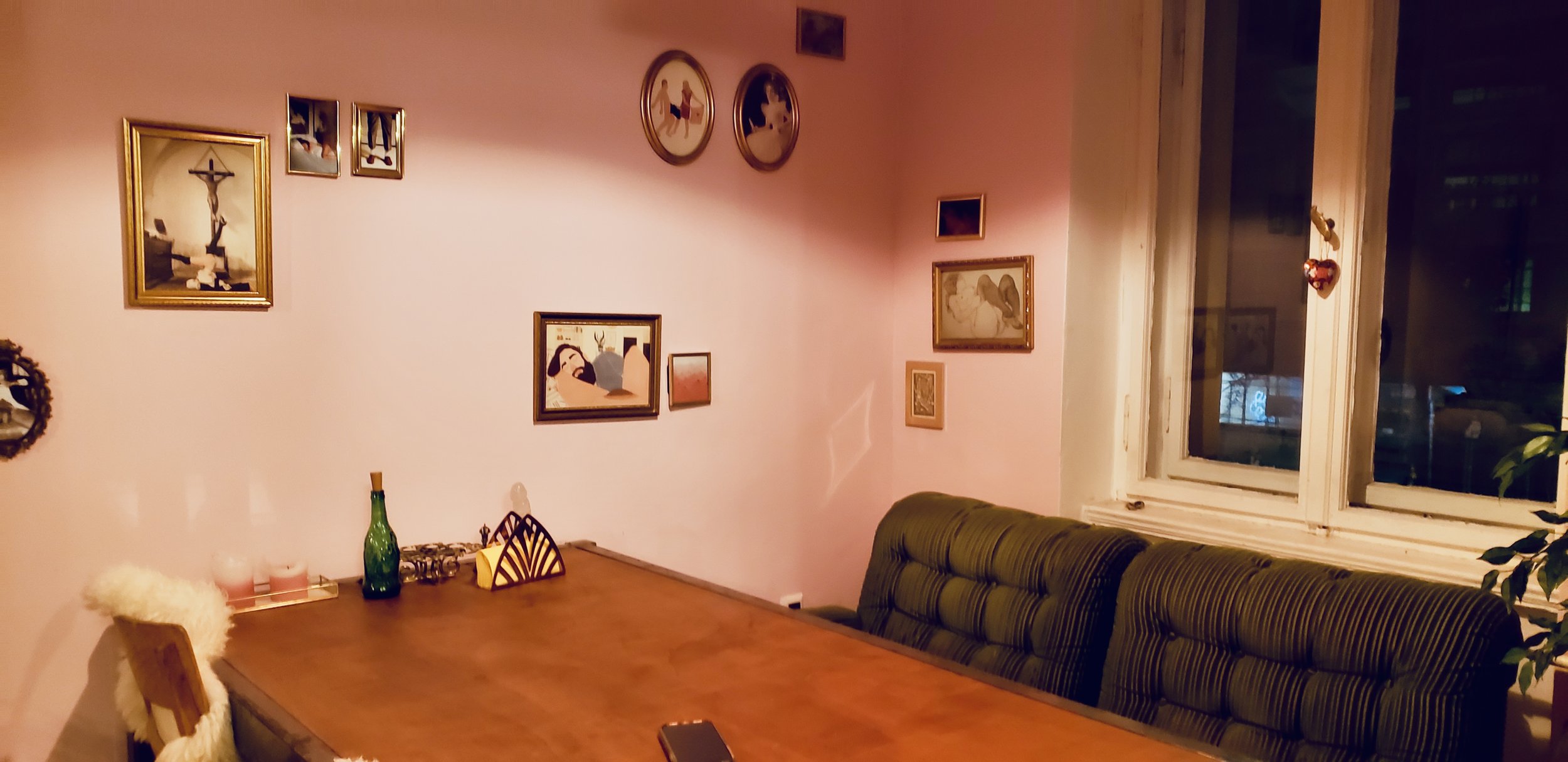 A cozy dining room with pink walls decorated with various framed pictures and artwork. There is a wooden dining table with a few items on it, including a green bottle and a yellow object. A cushioned bench runs along the wall under a window, with a green and black striped pattern. A small potted plant is visible near the window.