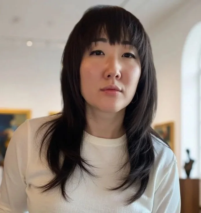 Portrait of a young woman with dark, shoulder-length hair and bangs, wearing a white shirt, standing indoors with art on the wall behind her.